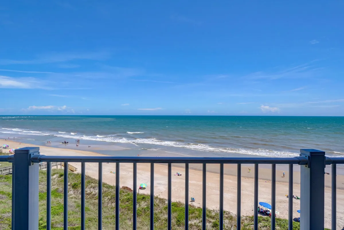 A panoramic view of the beach and ocean, with beachgoers enjoying the sand dunes under a blue sky. The scene captures the expansive Atlantic horizon and the vibrant, tranquil atmosphere of the coastal resort setting.