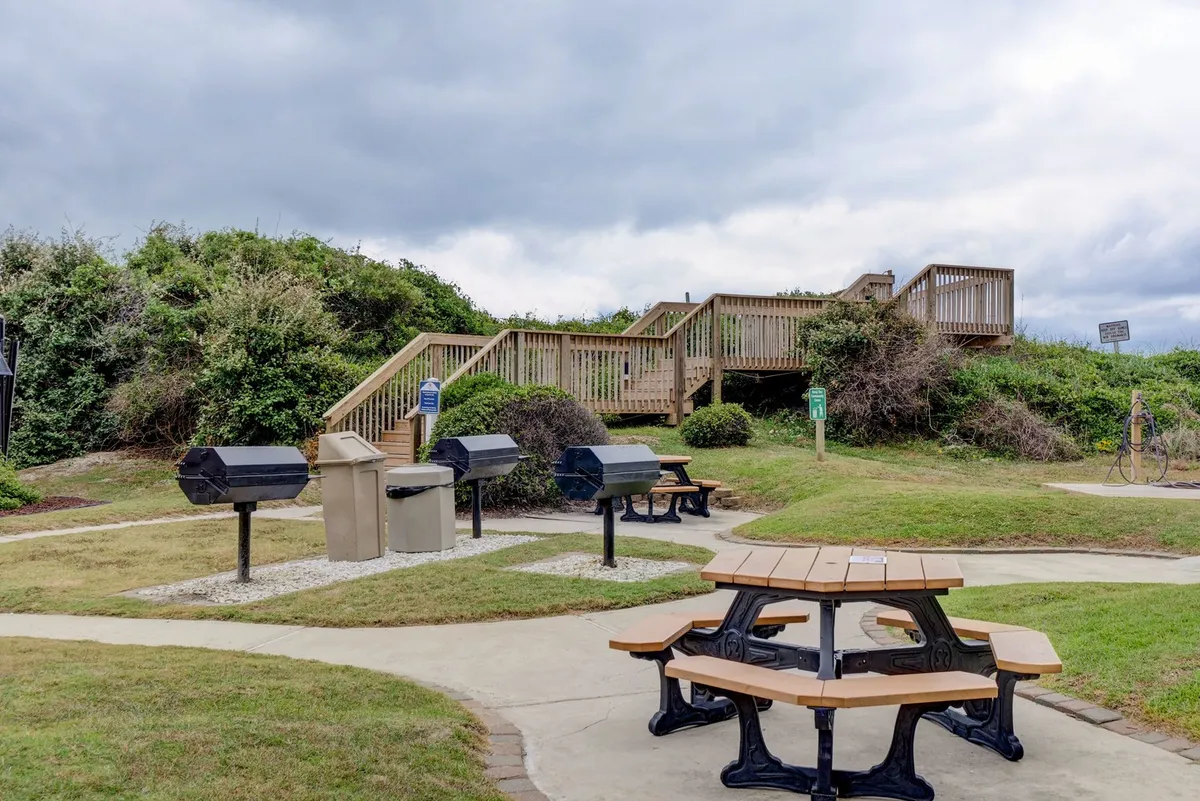 An outdoor communal area featuring picnic tables and several grills on a wooden deck. The atmosphere is friendly and perfect for barbecues, with plenty of space for socializing and enjoying meals outdoors.