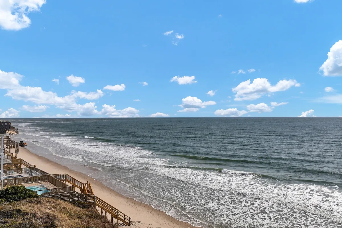 This hero image captures a sweeping beachfront scene at North Topsail Beach, with an expansive view of the ocean, a private wooden boardwalk leading directly to the sand, and the inviting shoreline stretching into the distance. The tranquil setting hints at activities like sunrise yoga, sunbathing, and evening bonfires, with the gentle waves and open sky creating a serene, inviting atmosphere.