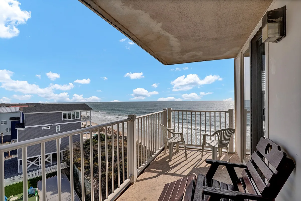 An exterior shot focused on a private balcony with a railing and seating area, set against a stunning ocean backdrop. The scene evokes relaxation, with the sun setting over the water and a tranquil mood ideal for morning coffee or evening toasts.