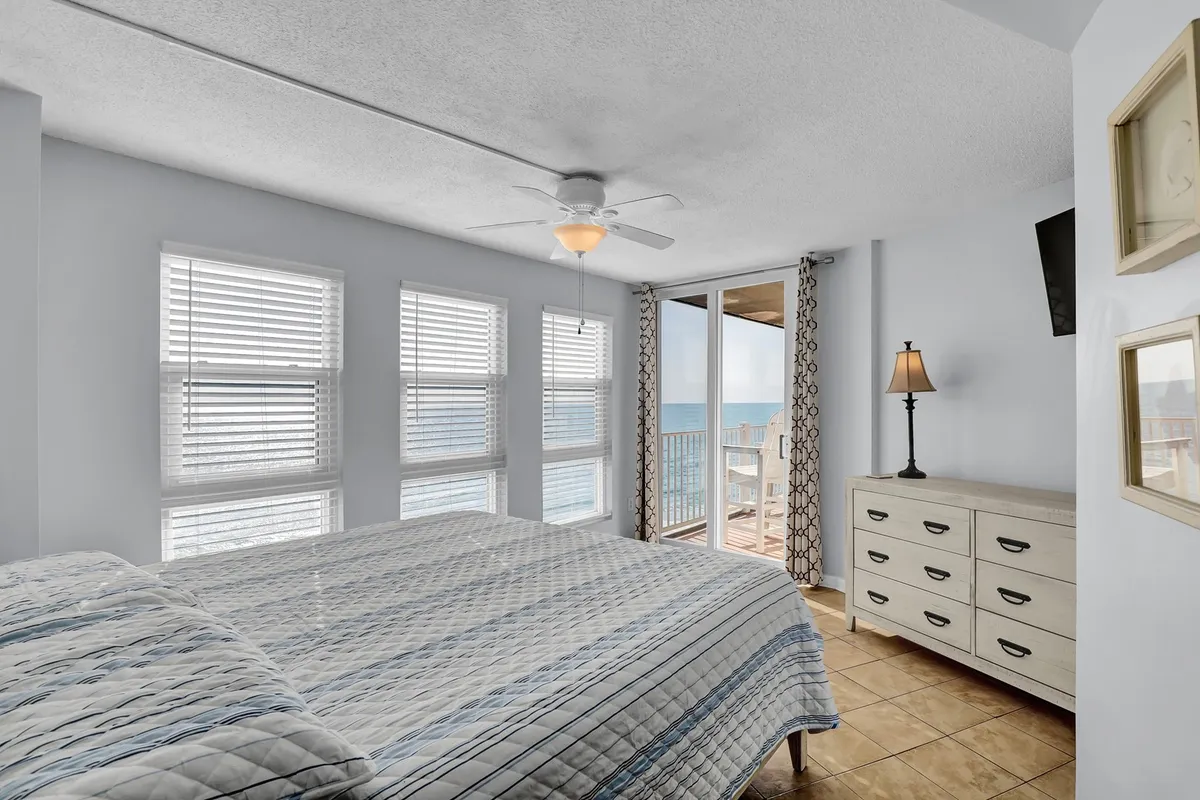 A serene bedroom with a comfortable bed, large windows, a sliding door offering access to a private balcony, a dresser, and a lamp. Natural light fills the room, emphasizing its peaceful and inviting atmosphere.