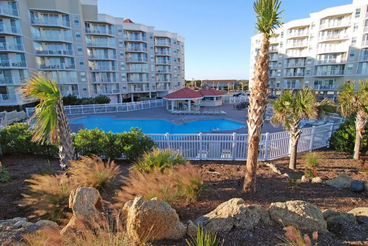 This image showcases a vibrant swimming pool surrounded by palm trees, adjacent buildings, and a poolside cabana. The setting exudes a resort feel with inviting blue waters, lush greenery, and ample space for relaxing or socializing poolside.