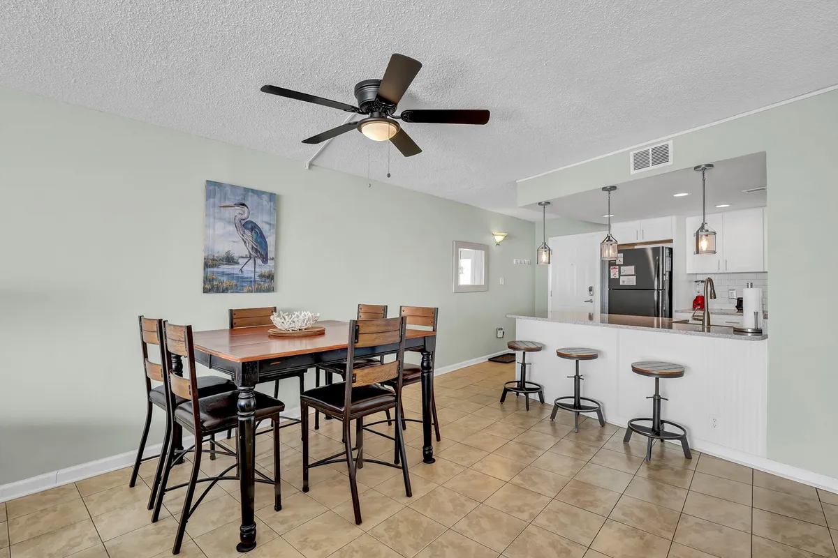 A bright, casual dining space with a dining table, bar stools at the kitchen island, and a wall painting of a bird. The kitchen area is open and inviting, suitable for both family dining and social gatherings.